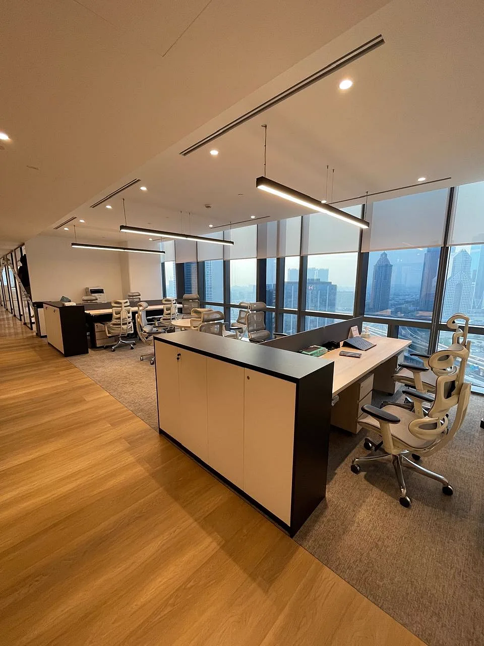 Open-plan workspace with linear suspended LED lighting, storage credenzas and ergonomic chairs beside floor-to-ceiling windows overlooking Dubai