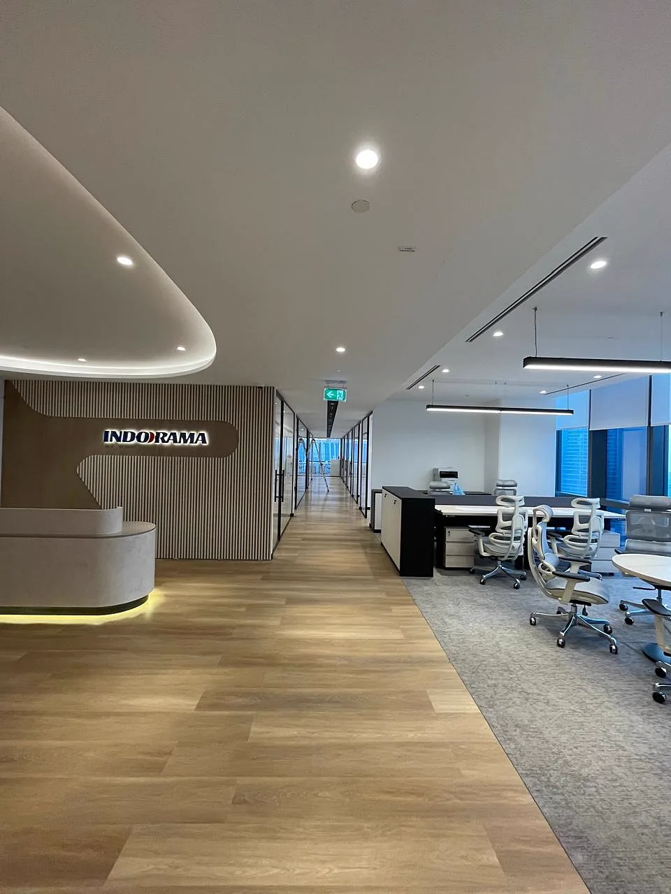 Completed Indorama corporate office with illuminated branding wall, curved reception desk, glass-partitioned corridor and open-plan workstations